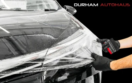 A professional installer wearing black gloves applies a transparent paint protection film (PPF) to the front of a black car at Durham Autohaus. The glossy surface is covered in water and soap solution to ensure smooth application, while the installer uses a squeegee for precise placement.