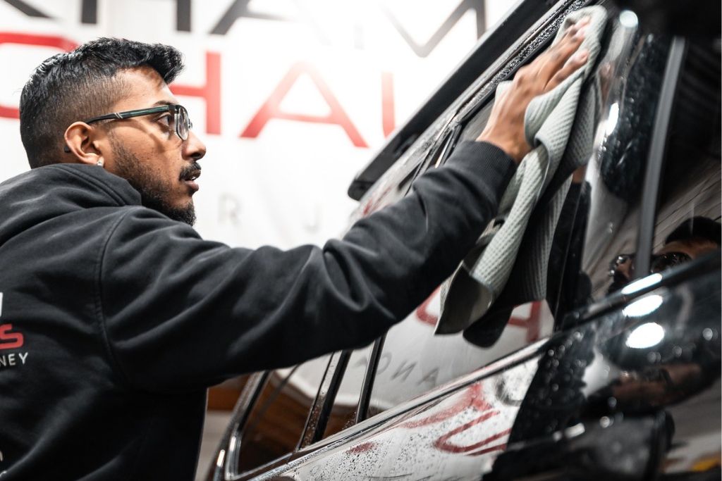 A professional installer wearing glasses and a black hoodie carefully wipes down a tinted car window with a microfiber cloth. The dark tint reflects the surroundings, while a large sign in the background hints at an automotive shop setting. The attention to detail highlights the protective benefits of window tint, including added safety in case of accidents.