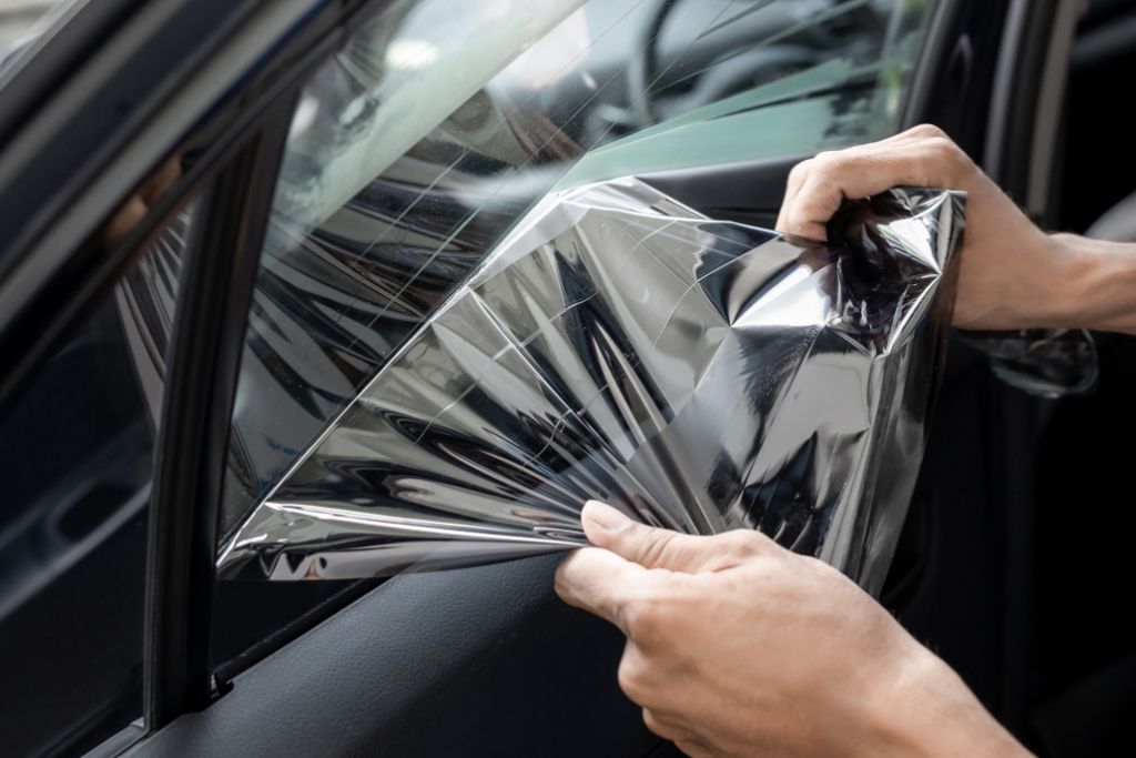 A person carefully peels a dyed window tint film from a car window, revealing the glass underneath. The semi-transparent film provides privacy and a sleek appearance but has a lighter tint compared to other options. The reflection on the film highlights its thin, flexible nature.