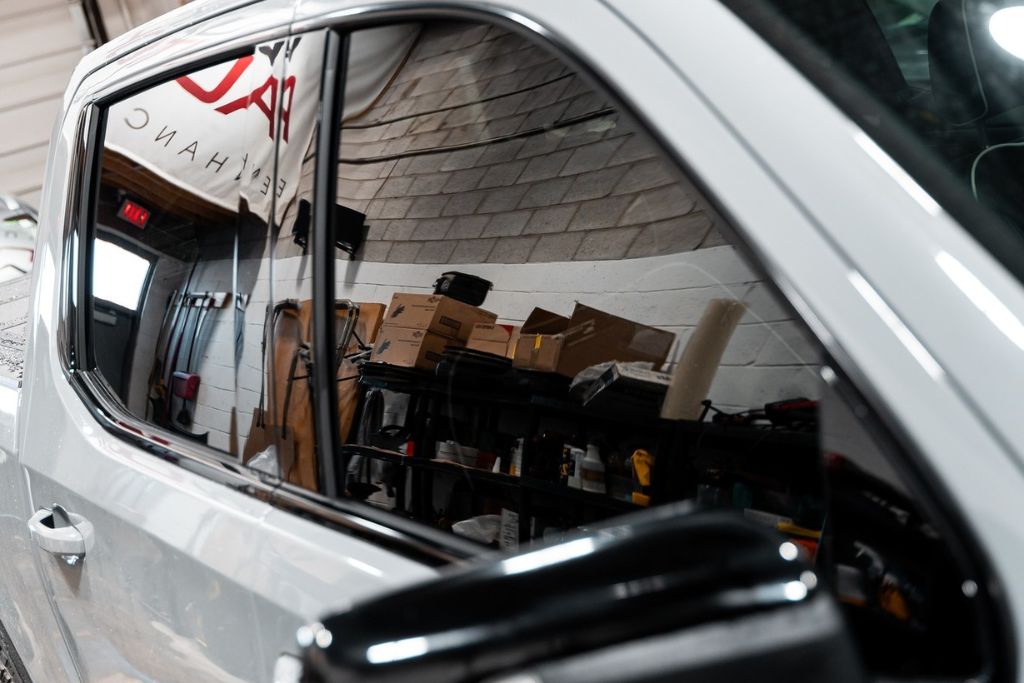 A close-up of a car's tinted window reflecting the interior of a garage with shelves stocked with boxes and tools. The dark tint reduces visibility into the vehicle, enhancing privacy and security.