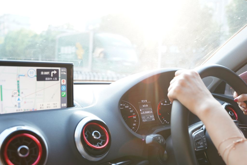 A driver grips the steering wheel while bright sunlight streams through the windshield, illuminating the dashboard and interior. The car’s navigation screen displays a map, while the air vents and speedometer are visible. The intense heat and glare suggest the benefits of heat reduction window tint to keep the cabin cooler and more comfortable.