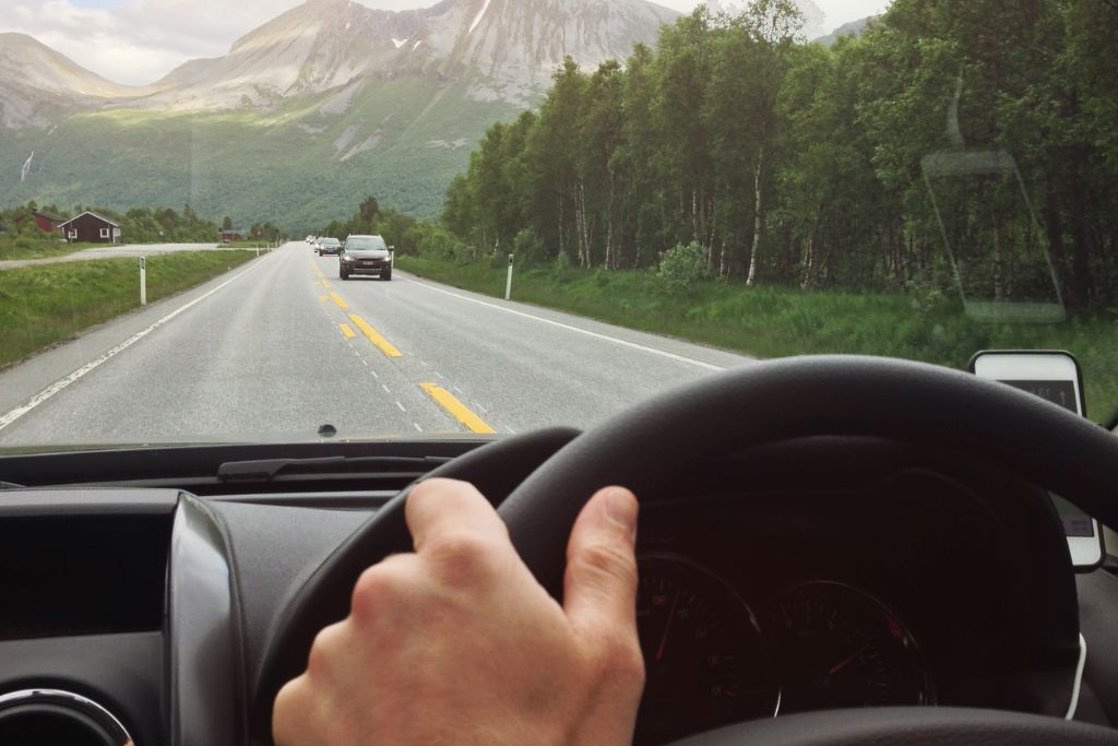 A driver grips the steering wheel while cruising down a scenic highway surrounded by lush greenery and towering mountains. The windshield provides a clear view of the road ahead, with minimal glare from natural light. The reduced reflections suggest the presence of glare-reducing window tint, enhancing visibility and driving safety.