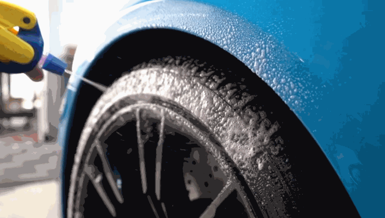 A vibrant blue sports car?s tire gets coated in cleaning foam as a detailer sprays it using a yellow and blue bottle, preparing the wheel for a deep clean.