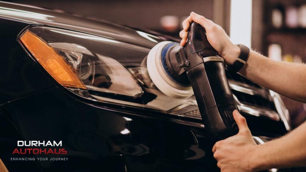 Close-up of a car headlight being polished with a power buffer by a technician, showcasing the detailing process at Durham Autohaus. The black vehicle?s surface reflects the surroundings, and the shop?s branding is visible in the corner.