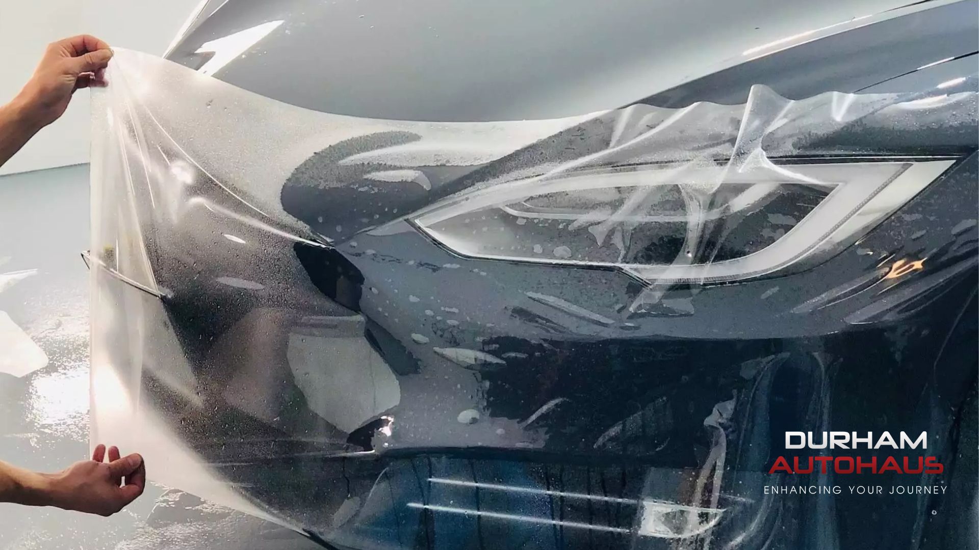 Hands pulling a clear paint protection film over a gray car?s headlight and front bumper, with the Durham Autohaus logo in the corner.