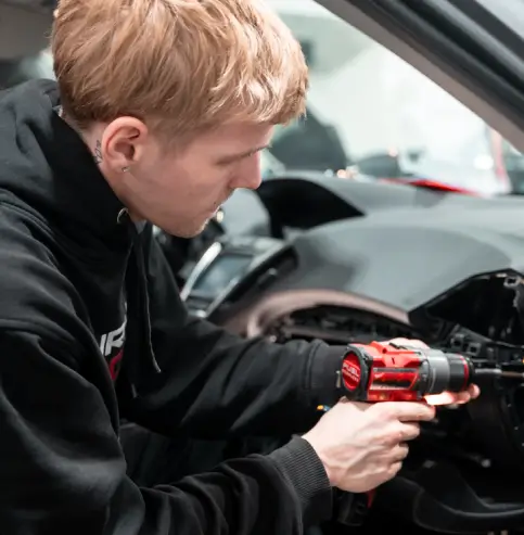 A young male technician with blond hair and a black t-shirt is working on the interior of a luxury car, reaching into the glove compartment or dashboard area. The car features a brown leather interior with wood trim and a sleek center console.